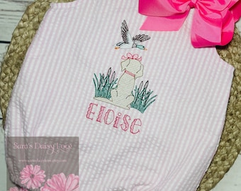 May include: A light pink and white seersucker bubble romper featuring a large hot pink hair bow at the shoulder. The front is embroidered with a tan dog, green reeds, a flying mallard duck, and the name Eloise in pink cursive lettering.