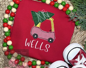 May include: A red t-shirt featuring an embroidered graphic of a car carrying a green Christmas tree on its roof. The car has a red and white checkered pattern. The name WELLS is embroidered in white below the car. A string of red, green, and cream wooden beads surrounds the shirt.