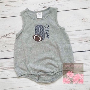 May include: Gray sleeveless baby onesie with a blue and white striped number "0" and a brown football. The onesie is personalized with the name "Oliver".