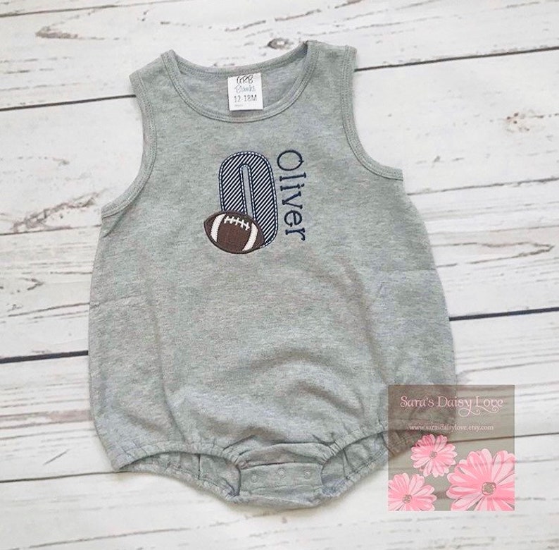 May include: Gray sleeveless baby onesie with a blue and white striped number "0" and a brown football. The onesie is personalized with the name "Oliver".