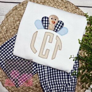 May include: White long-sleeved shirt with a turkey applique and a monogram. The turkey has blue and navy gingham feathers. The monogram is in a tan gingham pattern. The shirt is paired with navy and white gingham pants.