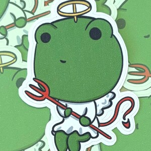 May include: A green frog sticker with a halo, wings, and a red pitchfork. The frog is wearing a white outfit and has a red tail. The background features similar stickers with a green and gold color scheme.