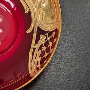 May include: A deep red glass saucer featuring an ornate gold-painted rim. The gold design includes swirling scrollwork and a delicate lattice pattern with small dots, creating a contrast against the translucent, vibrant red glass surface.