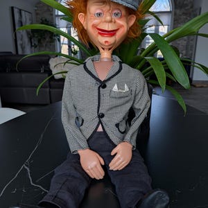 Può includere: Un pupazzo da ventriloquo vintage con un viso dipinto, capelli rossi e un berretto a scacchi. Indossa una giacca a scacchi bianca e nera, pantaloni scuri e scarpe nere. Il pupazzo è seduto di fronte a una pianta.
