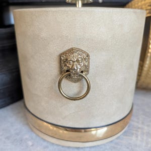 Könnte beinhalten: Ein zylindrischer Eiskübel mit beigefarbener wildlederartiger Oberfläche, der vorne einen goldfarbenen Metallgriff in Form eines Löwenkopfes mit Ring besitzt. Der Boden ist mit einem polierten goldfarbenen Metallband verziert und der Deckel hat einen kleinen goldenen Knopf.