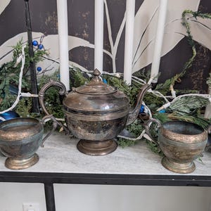 Op de afbeelding: Een antieke zilveren theeservies, bestaande uit een theepot en twee bijpassende kopjes, staat op een marmerachtig oppervlak. De set is aangetast en toont zijn leeftijd. De achtergrond omvat witte kaarsen, groen en een patroonachtergrond.