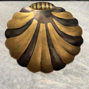 Peut inclure: Un plat décoratif en forme de coquillage avec des segments dorés et bronze foncé alternés. Le plat a un bord festonné et une petite poignée striée. La finition métallique lui donne un aspect vintage ou antique.