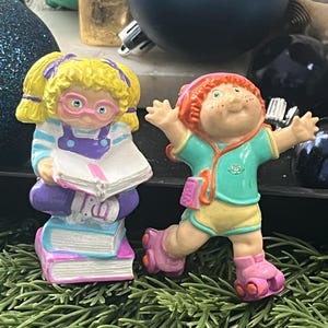 Könnte beinhalten: Zwei Vintage-Cabbage-Patch-Kids-Figuren. Eine ist ein Mädchen mit gelbem Haar, Brille und einem Buch. Die andere ist ein Mädchen mit rotem Haar, das Rollschuhe und ein türkisfarbenes Hemd trägt. Beide sind farbenfroh.