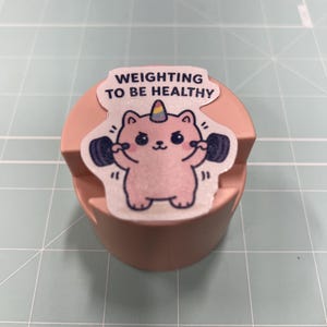 May include: A pink, cylindrical phone stand with a sticker featuring a cartoon cat lifting weights. The sticker reads "Weighting to be Healthy" and includes a rainbow unicorn horn.