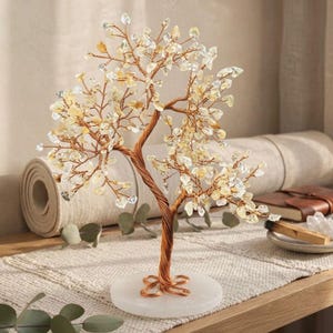 Puede incluir: Una escultura decorativa de árbol con un tronco y ramas de alambre de color cobre. Las ramas están adornadas con virutas de cristal transparentes y amarillas, que se asemejan a hojas. El árbol se asienta sobre una base blanca y redonda. La escultura se exhibe sobre una mesa de madera.
