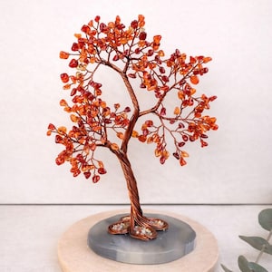 Peut inclure: Une sculpture artisanale en fil de fer représentant un arbre, avec un tronc et des branches cuivrées, ornées de feuilles de pierres précieuses orange et rouges. L'arbre repose sur une base en pierre grise, montée sur une plateforme en bois ronde.