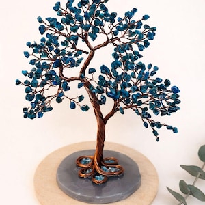 Könnte beinhalten: Handgefertigte Drahtbaumskulptur mit kupferfarbenem Stamm und Ästen, verziert mit leuchtend blauen Edelsteinblättern. Der Baum steht auf einem grauen Steinsockel, montiert auf einer Holzscheibe. Ein dekoratives Element für die Inneneinrichtung.