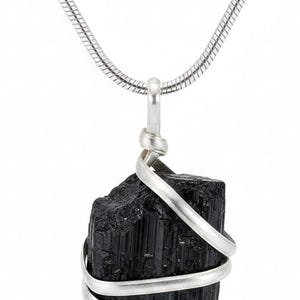 May include: A black tourmaline crystal pendant necklace. The raw, rectangular-shaped stone is wrapped with a silver-colored wire and hangs from a silver chain. The necklace is displayed against a white background.