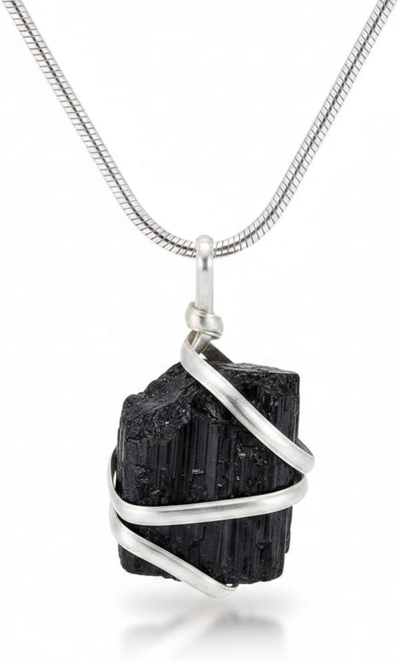 May include: A black tourmaline crystal pendant necklace. The raw, rectangular-shaped stone is wrapped with a silver-colored wire and hangs from a silver chain. The necklace is displayed against a white background.