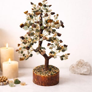 May include: A gemstone tree sculpture with a brown trunk and branches, decorated with multi-coloured stone chips. The tree is set in a round, wooden base. Two lit candles and crystals are in the background.