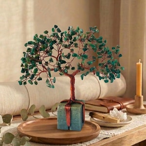 May include: A decorative tree sculpture with a copper wire trunk and branches, adorned with green gemstone leaves. The tree sits atop a teal-colored block, tied with a red ribbon, and is displayed on a wooden tray. A rolled-up yoga mat and a candle are in the background.
