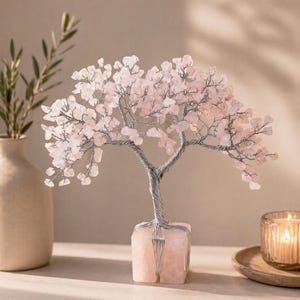 May include: A decorative tree sculpture with a silver wire trunk and branches, adorned with pink gemstone leaves. The tree is set in a square, light pink stone base. A candle in a glass holder and a vase with greenery are in the background.