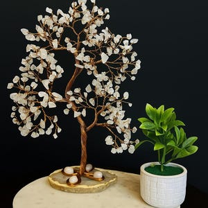 Puede incluir: Una escultura de árbol hecha a mano con alambre de cobre y hojas de piedras preciosas blancas, colocada sobre una base de piedra natural. Una pequeña planta de hojas verdes en una maceta blanca se encuentra junto al árbol sobre una superficie de mármol. El fondo es negro.