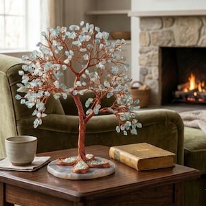 May include: A decorative tree sculpture featuring a copper wire trunk and branches adorned with numerous small, pale blue translucent stone chips. The tree is mounted on a circular, polished stone base with a smooth, light-colored surface.