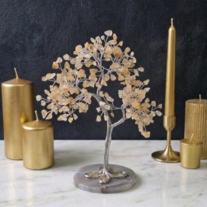 May include: A decorative tree sculpture with a silver-colored trunk and branches, adorned with amber-colored stones. The tree sits on a gray circular base. Several gold-colored candles of varying sizes are arranged around the tree, creating a warm, inviting ambiance.