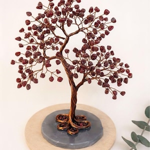 Könnte beinhalten: Eine handgefertigte Drahtbaumskulptur mit kupferfarbenem Stamm und Ästen. Die Äste sind mit kleinen, dunkelroten Edelsteinen geschmückt, die Blättern ähneln. Der Baum steht auf einem grauen Steinsockel, der auf einer hellen Holzbasis steht.