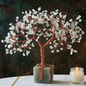 Könnte beinhalten: Eine dekorative Baumskulptur mit einem Stamm und Ästen aus Kupferdraht, verziert mit weißen Kristallblättern. Der Baum steht auf einem quadratischen, grauen Sockel. Eine brennende Kerze in einem Glas steht neben dem Baum.