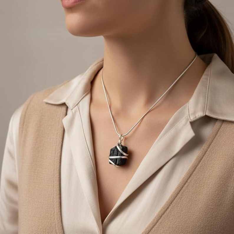 May include: A black tourmaline pendant necklace with a silver chain. The raw, dark gemstone is wrapped with silver wire. The necklace is worn over a cream-colored blouse and a tan vest.