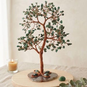May include: A decorative tree sculpture with a copper wire trunk and branches, adorned with green gemstone leaves. The tree sits on a grey stone base, displayed on a wooden surface. A lit candle in a glass holder is in the background.