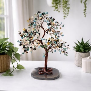 Könnte beinhalten: Eine dekorative Baumskulptur mit einem Stamm und Ästen aus Kupferdraht, verziert mit farbigen Edelstein-Chips. Der Baum steht auf einem grauen, oval geformten Steinsockel. Die Edelsteine sind in Blau-, Grün-, Gelb- und Brauntönen gehalten.