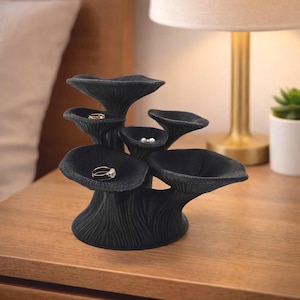 May include: A matte black jewelry organizer shaped like a cluster of mushrooms with multiple tiered caps. The structure features textured, ribbed stems and wide, concave tops that serve as small bowls for holding rings and small earrings.