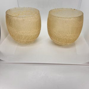 May include: Two small, round, light amber-colored glass cups. Each cup has a delicate, web-like pattern etched into the surface. The cups are displayed on a white surface.