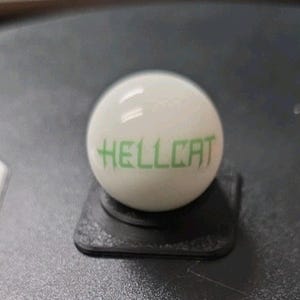 May include: A white billiard ball with the word "HELLCAT" printed in green. The ball is resting on a small, black, square stand. The background is a dark surface.