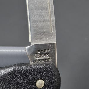 May include: A silver-colored metal knife blade featuring a stamped logo that reads CASE XX with five dots above and five dots below the text. The blade is attached to a textured black handle with a single visible silver metal rivet.