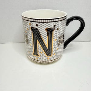 Op de afbeelding: Witte keramische mok met een zwart handvat. Het heeft een mozaïekpatroon met de letter "N" in zwart en goud. De mok heeft een honingraatontwerp en gouden accenten, met een zwarte rand aan de bovenkant.