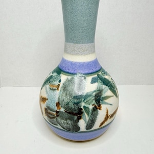 May include: A ceramic vase with a bulbous base and a slender neck. The vase features a light blue neck, a white and gray band, and a base with a blue and white floral design. The vase is approximately 8 inches tall.