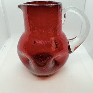 May include: A red glass pitcher featuring a distinct crackle texture throughout the body. The vessel has a rounded shape with a wide base, a narrow neck, a small pouring spout, and a clear, curved glass handle attached to the side.