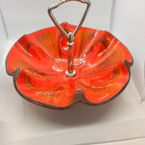 May include: A two-tiered serving dish with a vibrant orange and brown glaze. The dish has a scalloped edge and a silver-colored handle. The design is reminiscent of mid-century modern ceramics. The dish is perfect for serving appetizers or desserts.