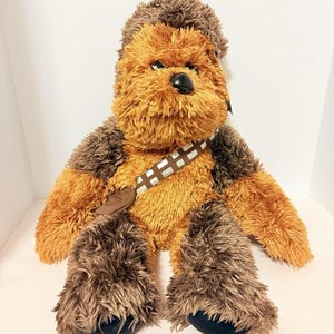 Op de afbeelding: Een Chewbacca pluche pop uit Star Wars. De pop heeft bruin en bruingeel haar, een zwarte neus en een bandolier. De pop zit met uitgestrekte armen en benen.