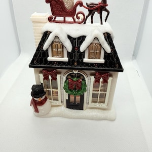 Könnte beinhalten: Dekorative Weihnachtshausfigur mit schwarzem Dach, weißen Schneebesätzen und einem Schneemann. Das Haus hat einen roten Schlitten und ein Rentier auf dem Dach. Die Fenster haben rote Schleifen und einen grünen Kranz.