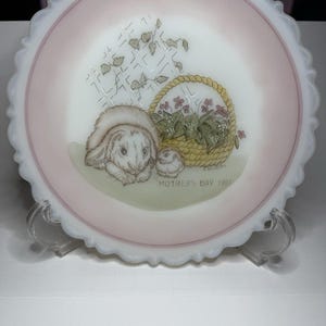 May include: A round, white milk glass plate with a scalloped edge and a soft pink border. The center features a detailed illustration of a rabbit and a small bird next to a woven basket of flowers, with the text MOTHER'S DAY 1991 printed below.