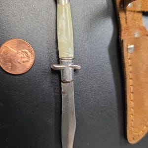 May include: A small, vintage knife with a silver-colored handle and blade, next to a penny and a brown leather sheath. The handle has a light-colored, pearlescent grip. The blade is silver and appears to be made of metal. The sheath is a light brown color.