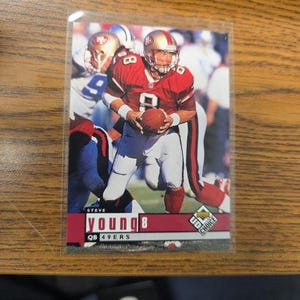 Op de afbeelding: Een Steve Young voetbalkaart met de quarterback in een rood en goud 49ers uniform, met een voetbal. De kaart heeft de tekst "STEVE YOUNG 8 QB 49ERS" en het UD Choice logo.