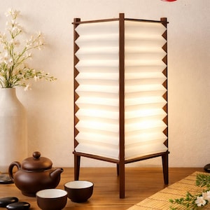 Può includere: Una lampada giapponese con struttura in legno marrone e paralume bianco a fisarmonica. La lampada è su un tavolo di legno, con una teiera, tazze e un vaso di fiori. Il testo "Japanese Lamp" è sopra la lampada.