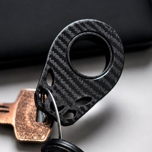 May include: A close-up of a set of keys, featuring a carbon fibre key ring and several metal keys. The key ring has a unique design with a circular hole. The keys vary in shape and size.