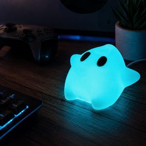 Puede incluir: Una l&aacute;mpara brillante de color azul claro con forma de fantasma y dos ojos oscuros de forma ovalada. La l&aacute;mpara emite una luz cian suave y vibrante, iluminando el &aacute;rea circundante con un brillo fr&iacute;o, creando un contraste con la oscuridad.