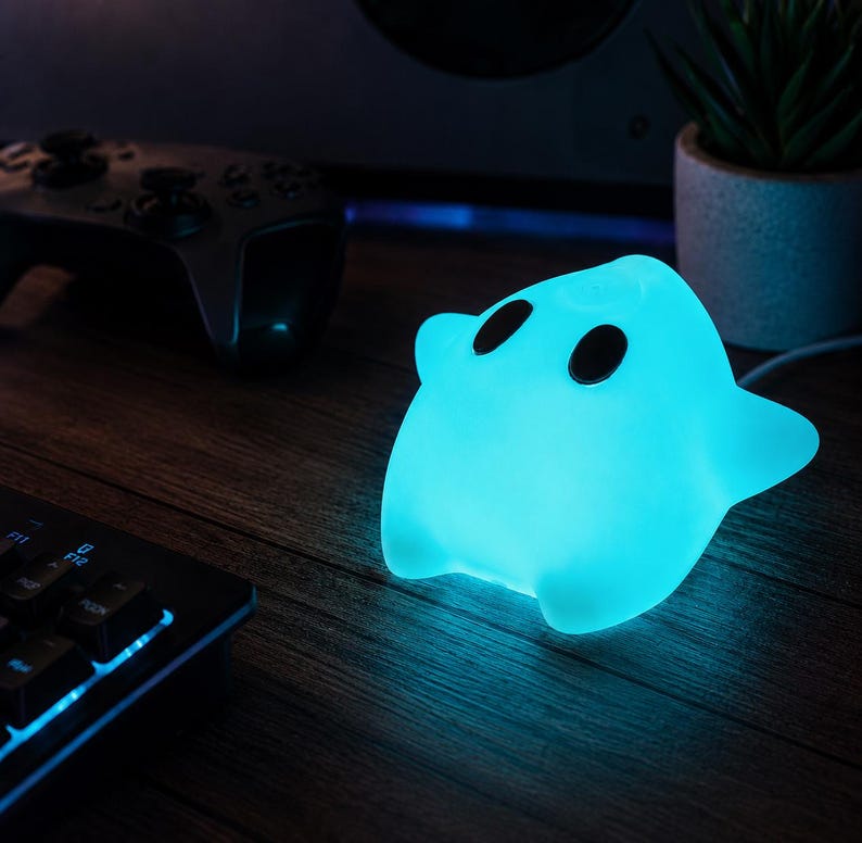 Puede incluir: Una l&aacute;mpara brillante de color azul claro con forma de fantasma y dos ojos oscuros de forma ovalada. La l&aacute;mpara emite una luz cian suave y vibrante, iluminando el &aacute;rea circundante con un brillo fr&iacute;o, creando un contraste con la oscuridad.