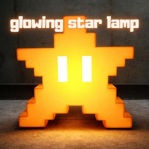 Pode incluir: Lâmpada estrela brilhante com design pixelizado. A lâmpada é laranja com dois olhos retangulares brancos e as palavras "glowing star lamp" em letras brancas acima. A lâmpada está sobre uma superfície cinza.