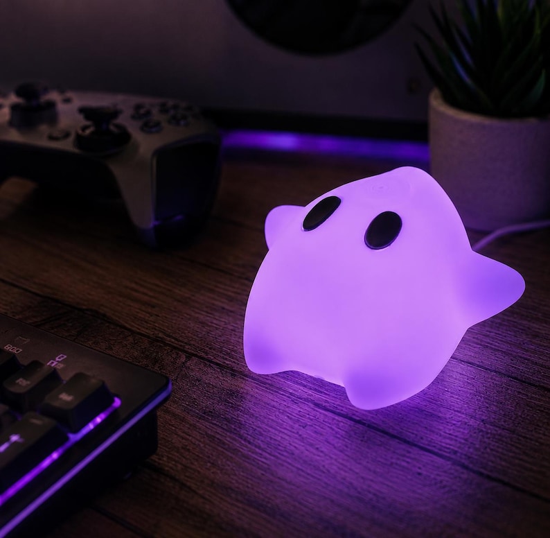 Puede incluir: Una l&aacute;mpara brillante de color morado con forma de estrella y dos ojos negros ovalados. La l&aacute;mpara emite una luz suave y vibrante que ilumina el &aacute;rea circundante. Presenta una superficie lisa y redondeada con un aspecto de personaje.