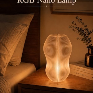May include: A glowing, ribbed, white RGB Nano Lamp sits on a wooden nightstand. The lamp has an hourglass shape and emits a warm light. The text "RGB Nano Lamp" is visible above the lamp. A bed and decorative items are in the background.