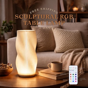Puede incluir: Una lámpara de mesa escultural RGB con un diseño ondulado único. La lámpara está hecha de un material de color claro y emite un brillo cálido. Tiene un control remoto para ajustar el brillo y el color. El texto en la imagen dice "Sculptural RGB Table Lamp".